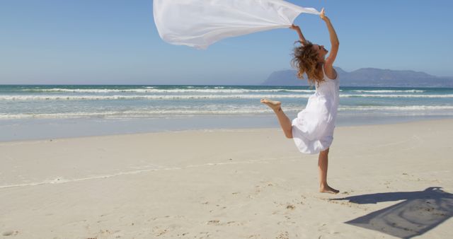 Woman in white dress dancing with white fabric on a sunny beach. Serene ocean and blue sky in background. Ideal for travel, lifestyle, and wellness themes, portraying freedom, joy, and relaxation.