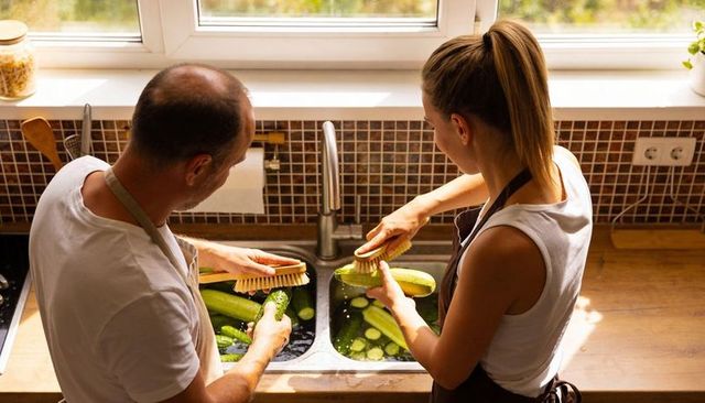 Couple washing cucumbers together at sunlit kitchen sink preparing fresh produce