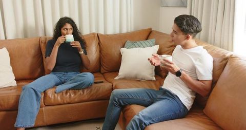 Young couple sipping coffee and talking on brown leather sofa in cozy modern living room