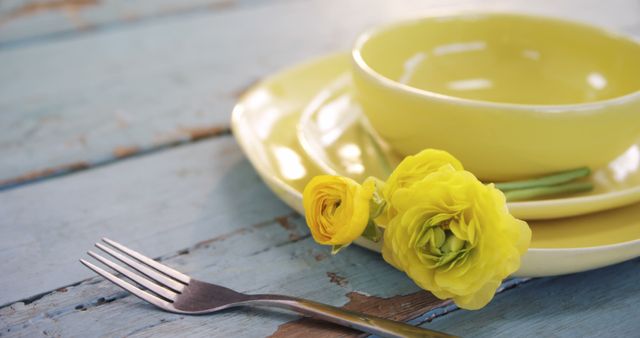 A yellow bowl and plate set accompanied by vibrant yellow flowers on a rustic blue wooden table, with copy space. The arrangement suggests a fresh, springtime setting ideal for a seasonal table setting or home decor theme.