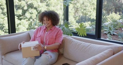 Happy Young Woman Opening Package on Modern Sofa in Bright Living Room