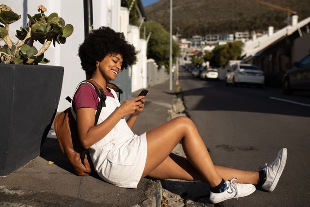 SIde view of a happy biracial woman enjoying free time in a city on a sunny day, using a smartphone, sitting on a pavement, wearing white mini dress and backpack.