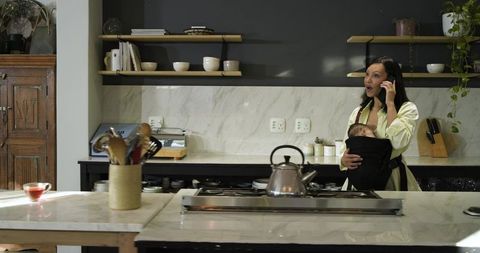 Hispanic mother multitasks in modern kitchen with baby in carrier