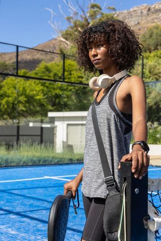 Athletic woman standing by padel net holding racket on vibrant blue court with headphones