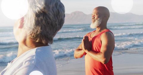 Mature couple practicing beach yoga at sunrise; man meditating in prayer pose