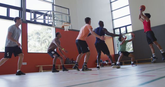 Group of men playing basketball in an indoor court. Men are wearing sports attire, some jumping and aiming for the basket. The high-ceiling court has large windows letting in natural light. This image can be perfect for sports event promotions, athletic apparel advertisements, fitness articles, and teamwork concepts.