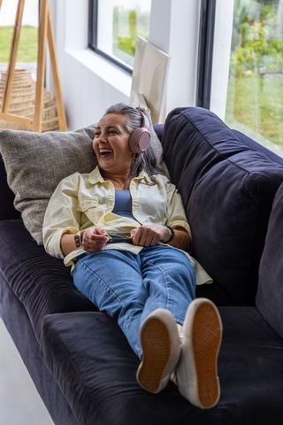 Senior woman relaxing on dark sofa wearing pink wireless headphones and enjoying music