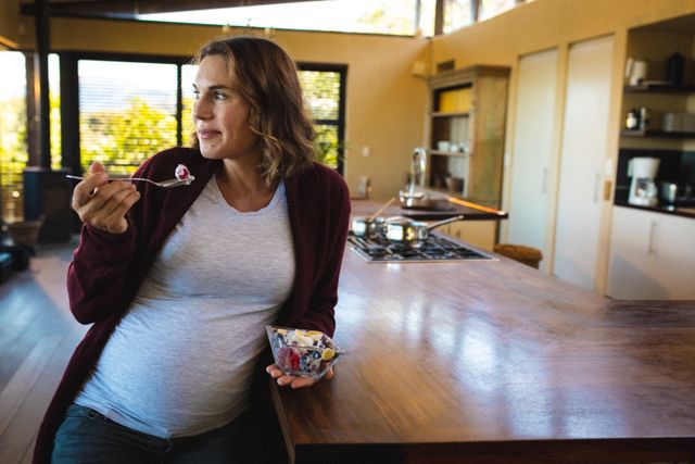 Pregnant woman savoring a nutritious breakfast at a kitchen island in a modern home. Ideal for use in articles or advertisements related to pregnancy, healthy eating, maternity lifestyle, and home living. Perfect for illustrating themes of anticipation, beginnings, and domestic life.