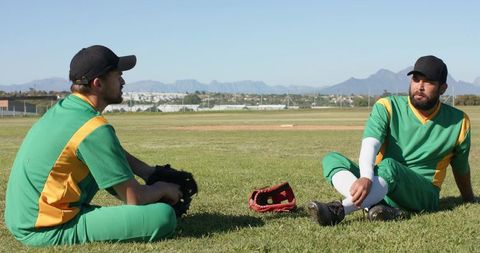 Baseball Teammates Sitting on Grass Talking Between Innings with Gloves and Mountain Backdrop