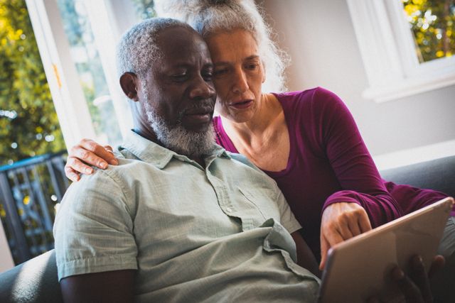 Senior couple sitting on couch in living room, using tablet together. Ideal for themes related to technology adoption among seniors, retirement lifestyle, bonding, and staying connected during quarantine or lockdown.