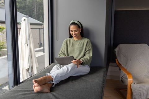 Asian woman relaxing on daybed by window using tablet in cozy minimalist interior