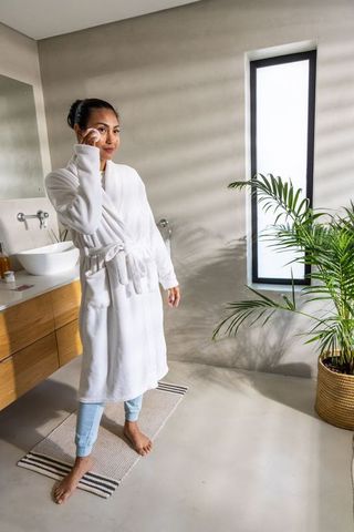 Asian woman applying facial pad in modern minimalist bathroom wearing white bathrobe