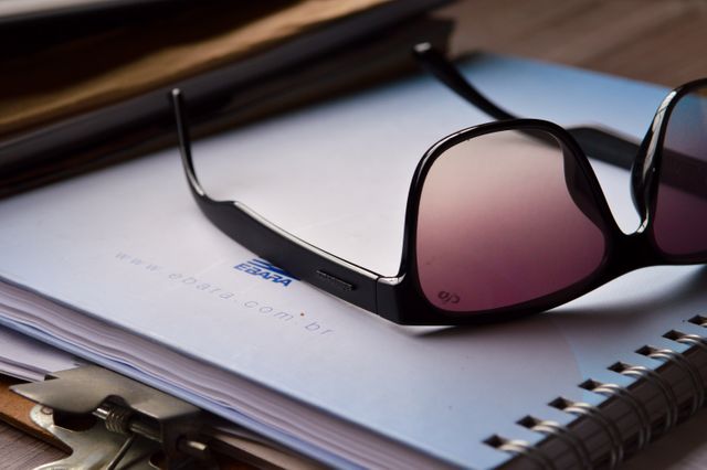 Stylish sunglasses lying on top of desk with documents and a notepad present. Ideal for illustrating office environments, business themes, work-from-home setups, fashion accessories, and organized workspaces.