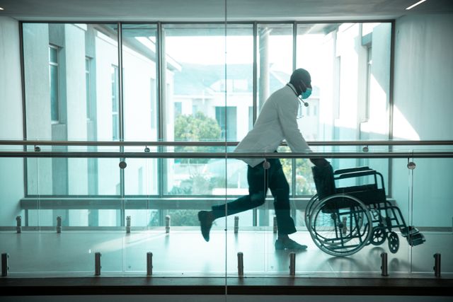 African american male doctor wearing face mask pushing wheelchair in hospital corridor. medical and healthcare services at hospital during covid 19 pandemic.