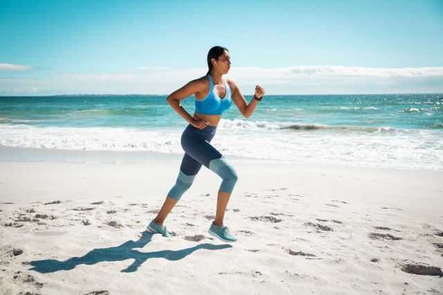 Biracial woman running on sandy beach wearing activewear and wireless earphones. Ocean waves in background. Ideal for promoting healthy lifestyle, fitness programs, outdoor activities, sportswear brands, and motivational content.