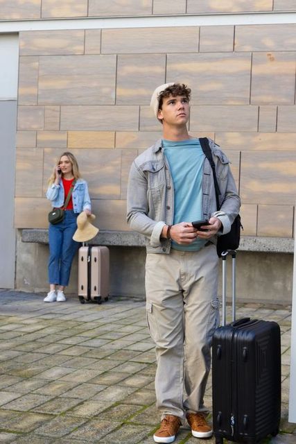 Young travelers waiting with rolling luggage and smartphones near modern urban building