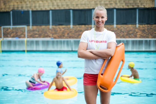 Confident female lifeguard standing poolside holding rescue can, smiling at camera. Children swimming and playing in the background. Ideal for use in articles about pool safety, lifeguard training, summer activities, and public pool facilities.