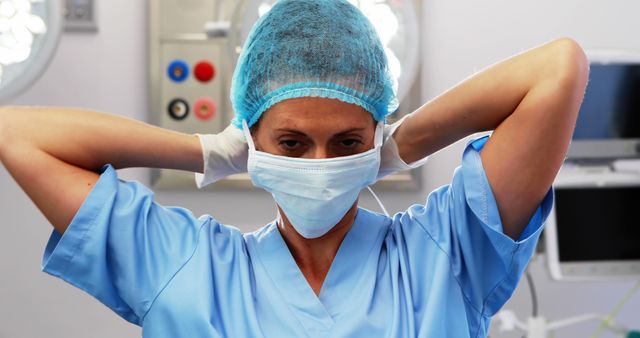 Female healthcare worker wearing surgical mask and scrub cap adjusting her protective gear. Represents themes of the medical profession, safety, and health care readiness. Suitable for articles about health care, posts about hospital life, and medical training materials.