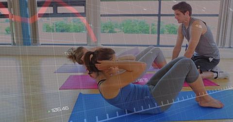 Woman performing abdominal crunches with trainer in sunlit fitness studio, group workout