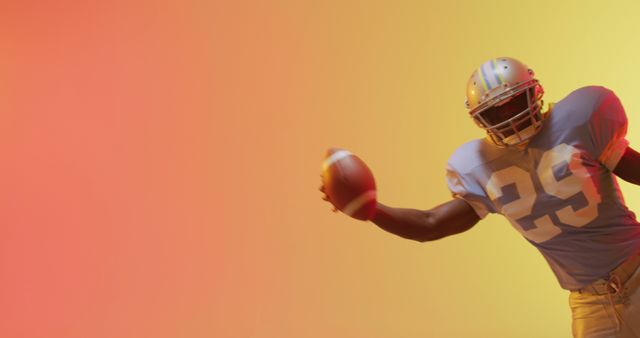 Football player holding a football while wearing helmet and uniform against a vibrant orange and yellow background. The dynamic pose emphasizes action and energy, making this suitable for use in sports promotions, team branding, and marketing materials for athletic events or products related to football.