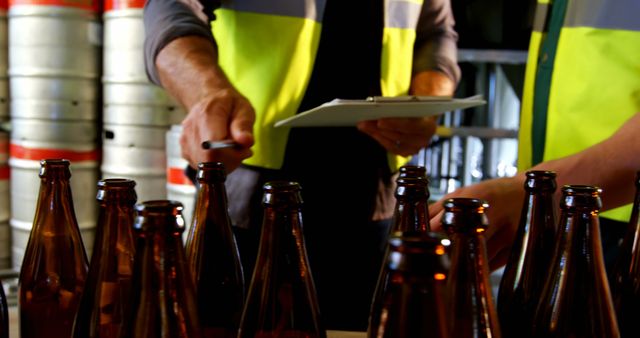 The male workers are inspecting empty beer bottles, noted down on a clipboard, ensuring quality control in a brewery. The photo is useful for topics like alcohol production processes, quality assurance in manufacturing, and workplace safety in production environments.