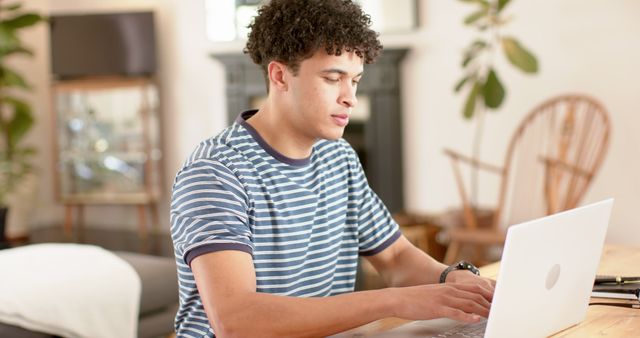 This young male, dressed in a casual striped shirt, is working at his laptop in a modern home office. Sunlight streams in, creating a pleasant atmosphere for remote work. This image is ideal for illustrating topics related to remote work, productivity, home offices, modern lifestyle, or youth engaged in professional tasks. It can be used for websites, articles, and marketing materials about remote work or tech use at home.