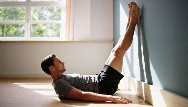 Man stretching legs on wall during post-workout cooldown at home in sunlit minimalist interior