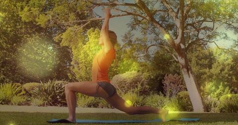 Woman practicing low lunge yoga on blue mat in sunny garden surrounded by lush trees