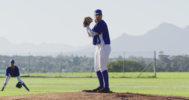 Baseball pitcher in blue-and-white uniform standing on mound with teammate in background on sunny day.Suitable for sports-related content, teamwork themes, baseball promotions, and athletic endeavors.