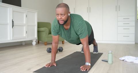 Black man performing high plank on yoga mat in sunlit living room with dumbbells