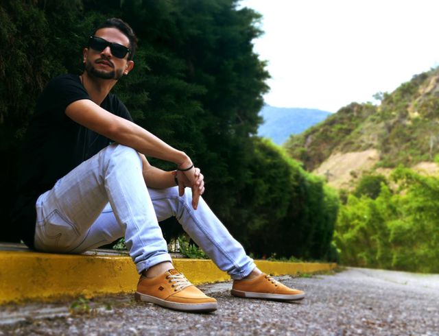 Young man wearing sunglasses and casual clothing, sitting on the curb in a natural, green environment with hills in the background. Perfect for promoting lifestyle brands, casual fashion, outdoor activities, and relaxed living.
