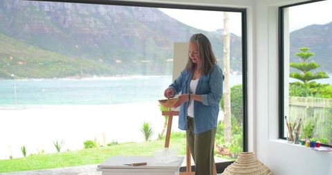 Mature female artist mixing paints in bright coastal studio with ocean view