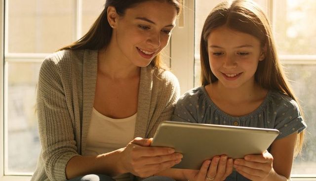 Mother and daughter sharing tablet by sunlit window, smiling, family bonding at home