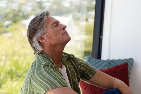 Older man relaxing by large window in sunlit living room, serene contemplative moment