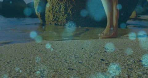Wading barefoot on pebbly beach with teal bokeh and rocky shoreline, summer calm