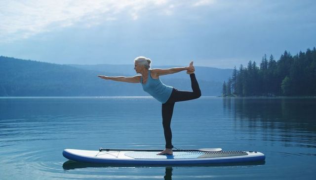 Senior woman practicing paddleboard yoga dancer pose on calm forest lake at sunrise