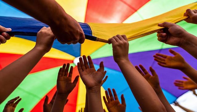 Children lifting rainbow parachute canopy reaching hands teamwork outdoor playground play