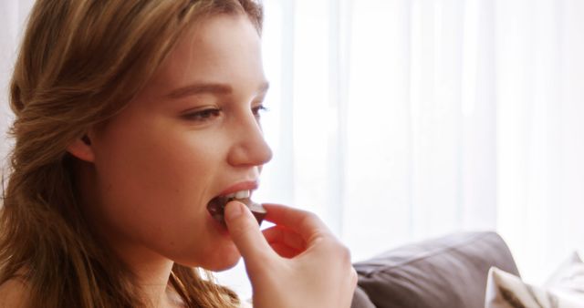 This image features a young woman enjoying chocolate while sitting in a casual environment. Ideal for promoting indulgence, lifestyle, and relaxation themes in advertising or social media content relating to food, self-care, and leisure.