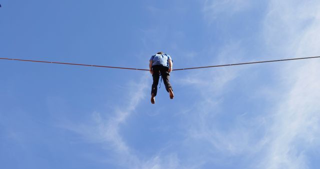 The image shows a tightrope walker balancing high above the ground on a tightrope with a clear blue sky as the backdrop. Perfect for themes of adventure, courage, balance, and skill. Could be used in articles, advertisements, or materials related to extreme sports, motivation, or overcoming challenges.