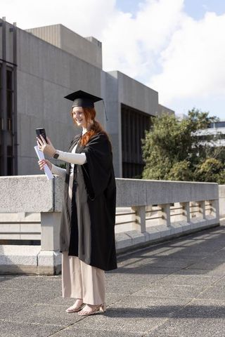 Smiling graduate taking selfie on university terrace in gown and cap holding diploma