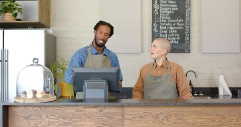 Diverse baristas collaborating at coffee counter using pos with cupcakes and chalkboard menu