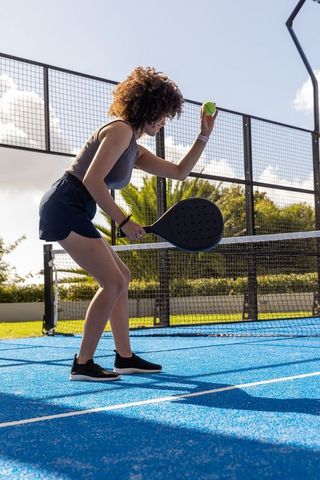 African american woman serving paddle tennis on bright blue court, athletic outdoor action