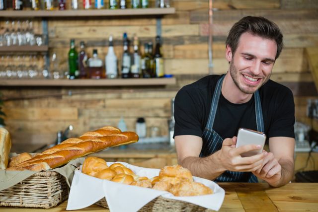 Waiter in a casual cafe setting using a mobile phone while standing behind a counter filled with bread and pastries. Ideal for use in articles or advertisements related to hospitality, customer service, technology in the workplace, or small business operations.