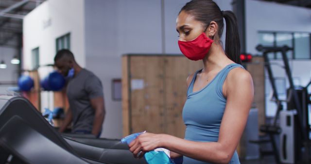 Woman wears red mask and disinfects treadmill in gym, emphasizing hygiene and safety during workout. Useful for illustrating health awareness, cleanliness in gyms, pandemic related safety protocols, and fitness environments.