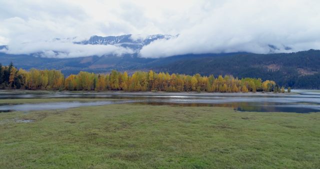 Feature idyllic autumn landscape capturing the beauty of a serene lake surrounded by colorful deciduous trees and distant mountains shrouded in clouds. Perfect for use in nature documentaries, travel blogs, outdoor adventure promotions, and landscape photography collections.