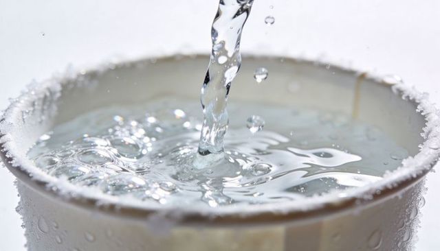 Pouring clear liquid into paper cup with crystalline rim macro water splash closeup