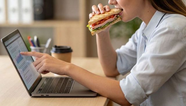 Woman eating sandwich while pointing at laptop charts during workday coffee break