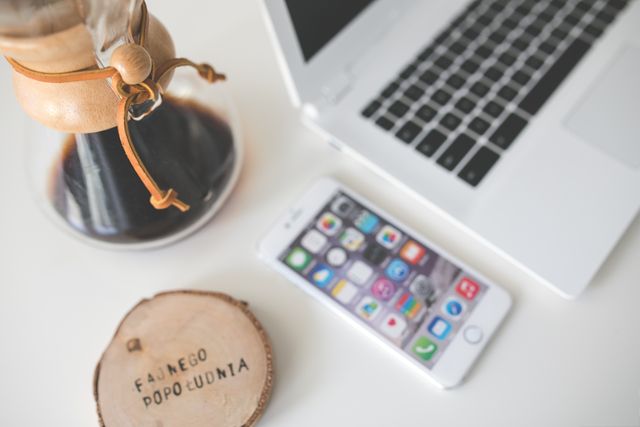 Ideal for showcasing remote work or home office setups, this image features a Chemex coffee maker, iPhone, and laptop on a white desk. Great for articles or advertisements relating to modern work environments, productivity, or technology.