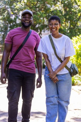 African american couple walking and laughing on sunlit park path with crossbody bags and cap