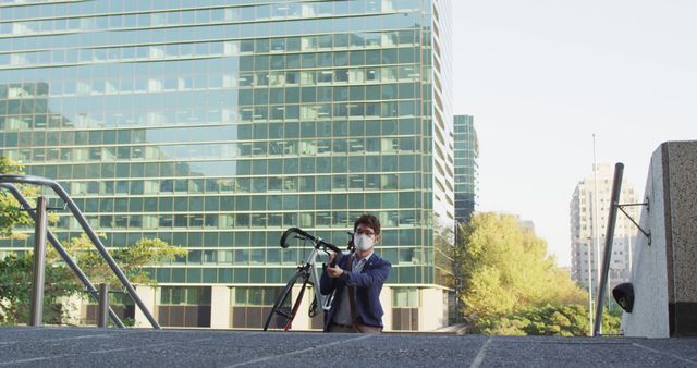Businessman wearing a face mask holding a bicycle, standing in front of a modern office building. Ideal for use in articles about urban living, safe commuting during the pandemic, transportation, eco-friendly travel options, and professional lifestyles in modern cities.